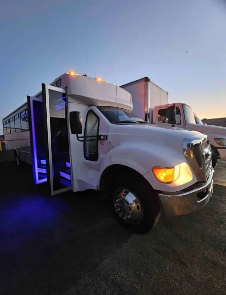 A white shuttle bus parked outdoors at dusk with its passenger door open, revealing blue LED lighting on the entry steps.