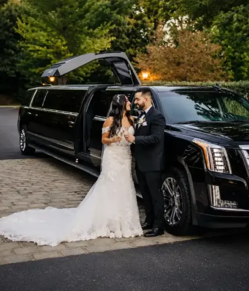 A bride and groom standing next to a black stretch limousine with the door open.