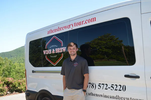 A man in a branded polo shirt standing in front of a white passenger van with 'Athens Wine & Brew Tours' logo and contact information.