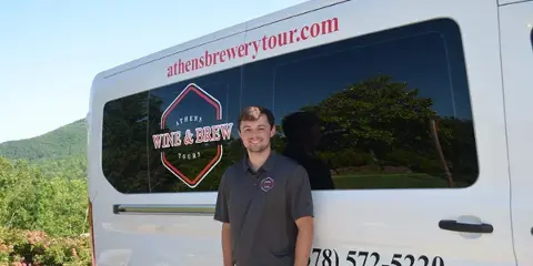 A man in a branded polo shirt standing in front of a white passenger van with 'Athens Wine & Brew Tours' logo and contact information.