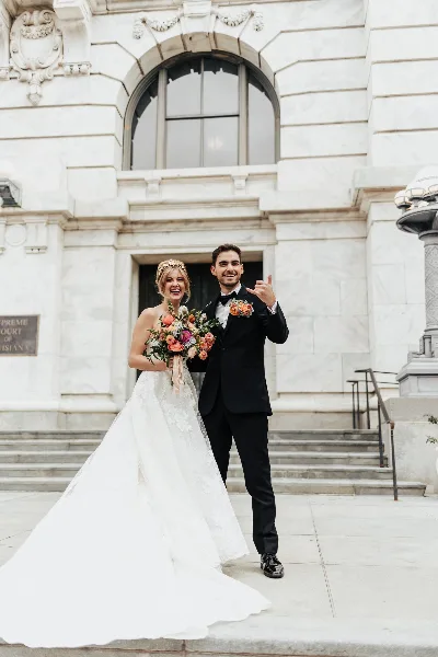 A bride in a white lace gown and a groom in a black tuxedo posing together in front of a stone building.