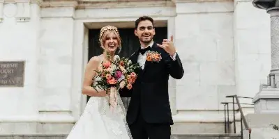 A bride in a white lace gown and a groom in a black tuxedo posing together in front of a stone building.