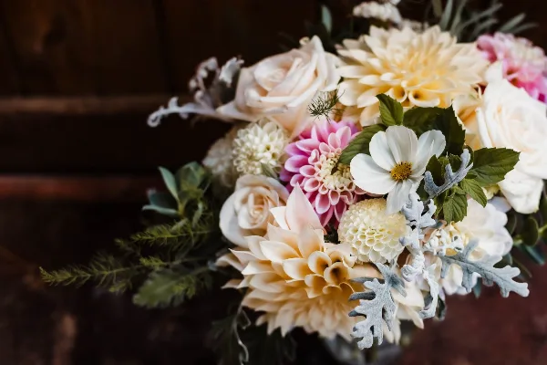 A close-up of a wedding floral arrangement featuring cream roses, yellow and pink dahlias, and dusty miller foliage.