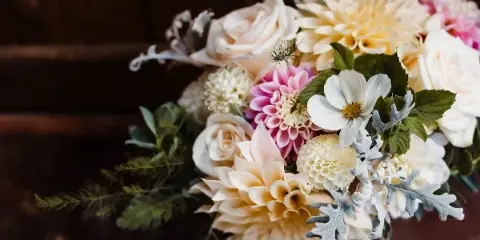 A close-up of a wedding floral arrangement featuring cream roses, yellow and pink dahlias, and dusty miller foliage.