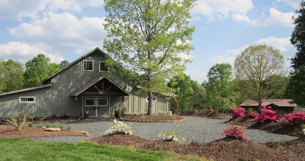 Exterior view of a large grey barn with a gravel courtyard, trees, and flowering bushes.