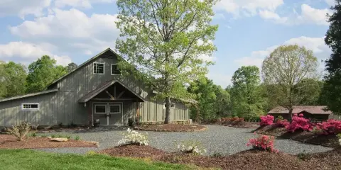 Exterior view of a large grey barn with a gravel courtyard, trees, and flowering bushes.