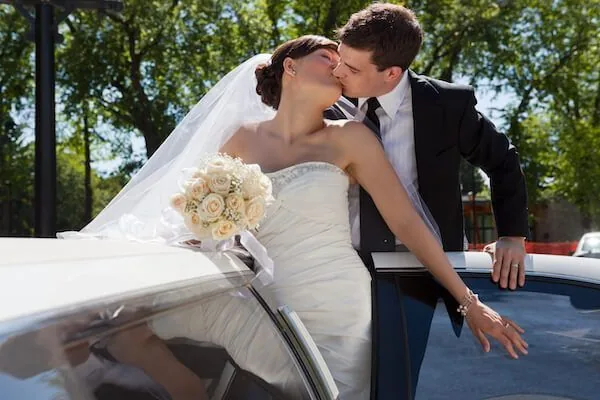 A bride and groom kissing next to a white limousine.