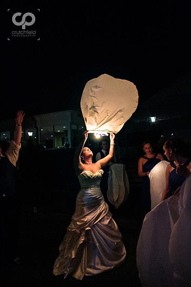 A bride in a formal gown releases a glowing sky lantern during a night event.