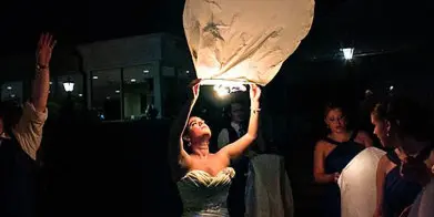 A bride in a formal gown releases a glowing sky lantern during a night event.