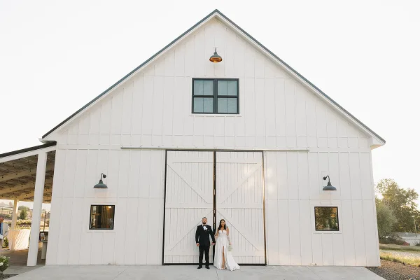 Exterior view of a large white barn with a couple standing in front of the main doors.
