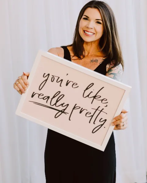A smiling woman with long brown hair and a black top holds a framed sign that reads 'you're like, really pretty.'