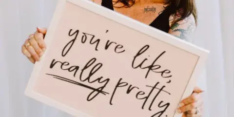 A smiling woman with long brown hair and a black top holds a framed sign that reads 'you're like, really pretty.'