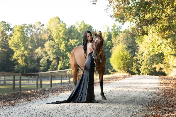 A professional portrait photograph of a woman in a long black dress standing next to a brown horse on a gravel path surrounded by trees.