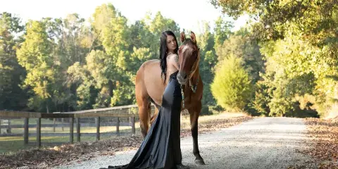 A professional portrait photograph of a woman in a long black dress standing next to a brown horse on a gravel path surrounded by trees.