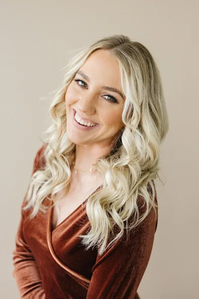 A smiling blonde woman in a brown velvet top, photographed in a studio setting.