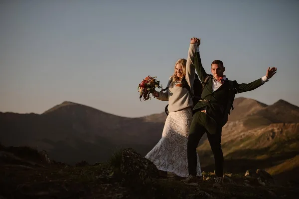 A bride and groom holding hands and raising their arms in celebration on a mountain peak with a bouquet and backpacks.