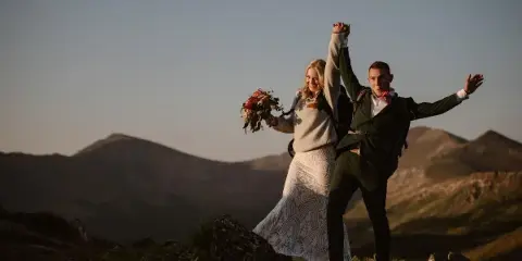 A bride and groom holding hands and raising their arms in celebration on a mountain peak with a bouquet and backpacks.