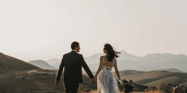 A bride and groom walking hand-in-hand through a scenic hilly landscape at sunset.