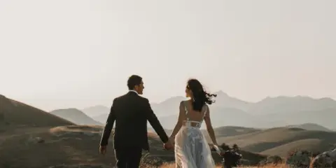 A bride and groom walking hand-in-hand through a scenic hilly landscape at sunset.