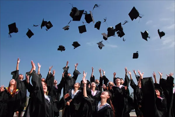 A group of graduates in black gowns throwing their caps into the air against a blue sky.