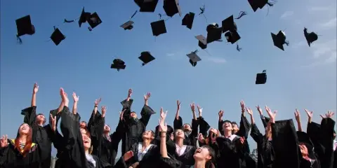 A group of graduates in black gowns throwing their caps into the air against a blue sky.