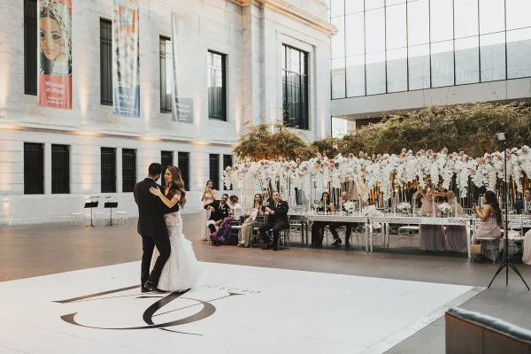 A bride and groom dancing at a wedding reception in a large venue with a long, flower-decorated head table and seated guests.