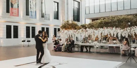 A bride and groom dancing at a wedding reception in a large venue with a long, flower-decorated head table and seated guests.