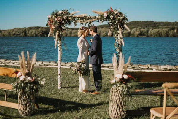 A bride and groom standing under a floral arch at a lakeside wedding venue.