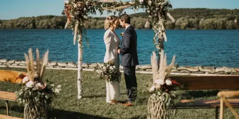 A bride and groom standing under a floral arch at a lakeside wedding venue.