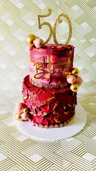 A two-tiered custom cake with red and pink frosting, gold leaf accents, and a 'Happy Birthday 50' topper.