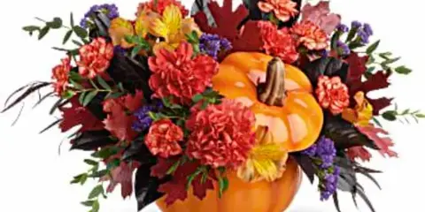 Autumn floral arrangement with red and orange carnations, alstroemeria, purple filler flowers, and dark foliage arranged around a ceramic pumpkin.
