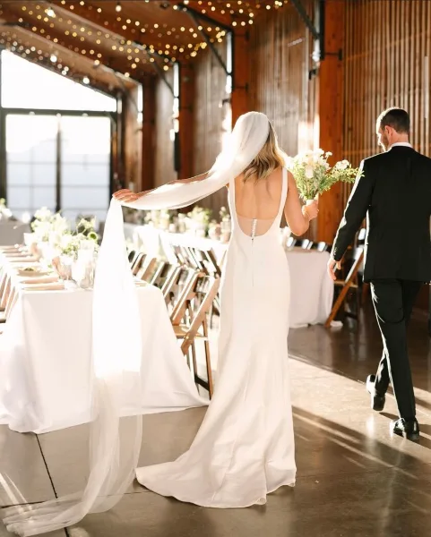 A bride and groom walking down an aisle in a rustic venue with long tables, white linens, and string lighting.