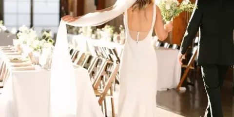 A bride and groom walking down an aisle in a rustic venue with long tables, white linens, and string lighting.