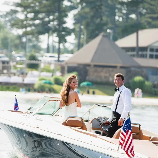 A bride and groom in formal wedding attire standing on a motorboat on a lake with a clubhouse in the background.