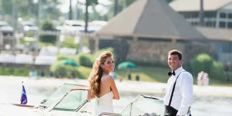 A bride and groom in formal wedding attire standing on a motorboat on a lake with a clubhouse in the background.