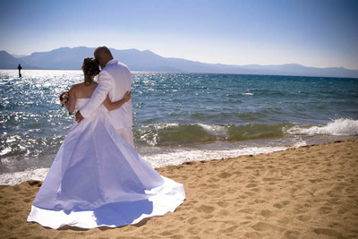 A bride and groom embracing on a sandy beach with the ocean in the background.