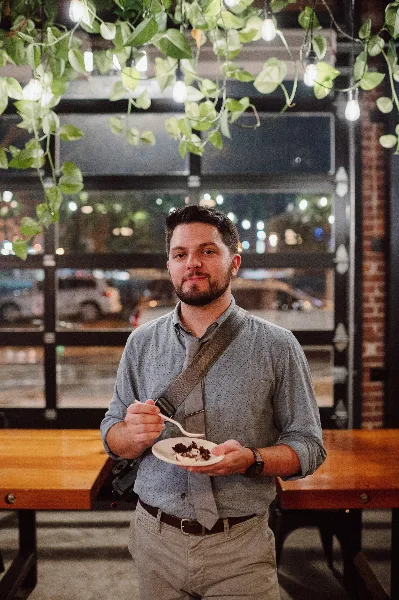 Portrait of a man with a beard wearing a grey shirt and tie, holding a camera and a plate of food in a restaurant setting.