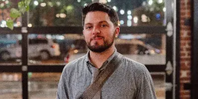 Portrait of a man with a beard wearing a grey shirt and tie, holding a camera and a plate of food in a restaurant setting.