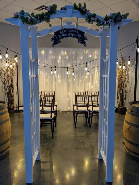 A wedding ceremony setup featuring a white arch decorated with greenery, a backdrop of white drapery with string lights, and rows of wooden chairs.