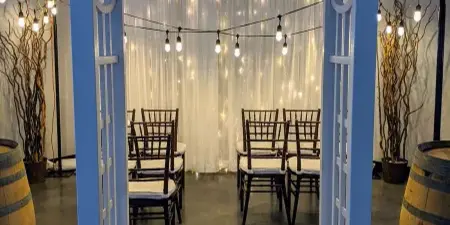 A wedding ceremony setup featuring a white arch decorated with greenery, a backdrop of white drapery with string lights, and rows of wooden chairs.
