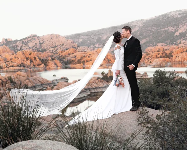 A bride and groom kissing on a rocky outcrop with a scenic desert landscape and water in the background.