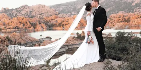 A bride and groom kissing on a rocky outcrop with a scenic desert landscape and water in the background.