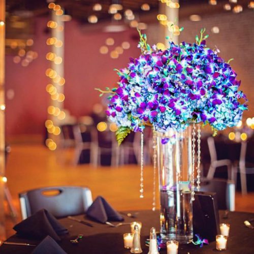 Tall blue and purple floral centerpiece in a glass vase on a reception table with candles and chairs in the background.
