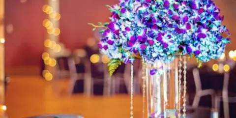 Tall blue and purple floral centerpiece in a glass vase on a reception table with candles and chairs in the background.