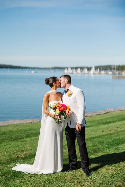 A bride and groom sharing a kiss on a grassy lawn with a lake and sailboats in the background.