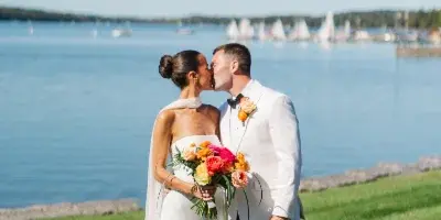 A bride and groom sharing a kiss on a grassy lawn with a lake and sailboats in the background.