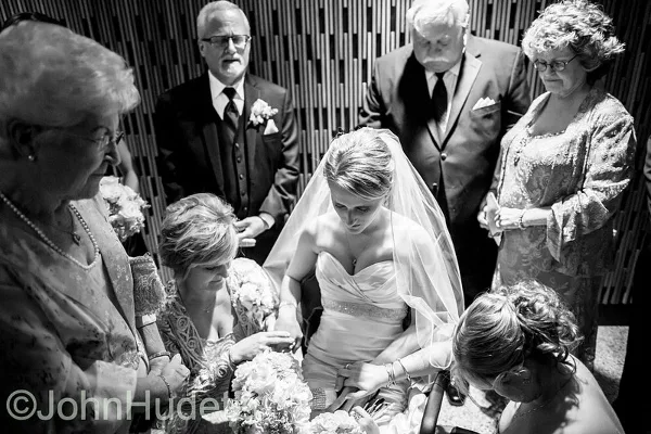 A black and white photograph of a bride in a wedding dress and veil, surrounded by family members, including a woman holding a bouquet of flowers.