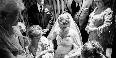 A black and white photograph of a bride in a wedding dress and veil, surrounded by family members, including a woman holding a bouquet of flowers.
