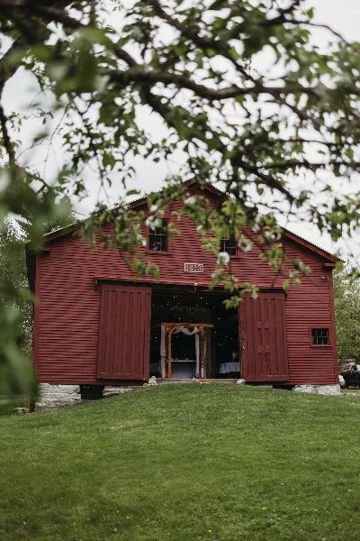 Exterior view of a red barn with the number 1830 on the front, set on a grassy lawn with trees in the foreground.