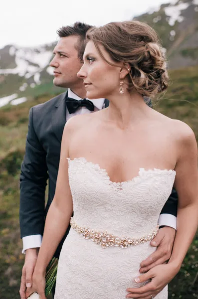 A bride in a white lace dress and a groom in a tuxedo standing together outdoors with a mountainous background.